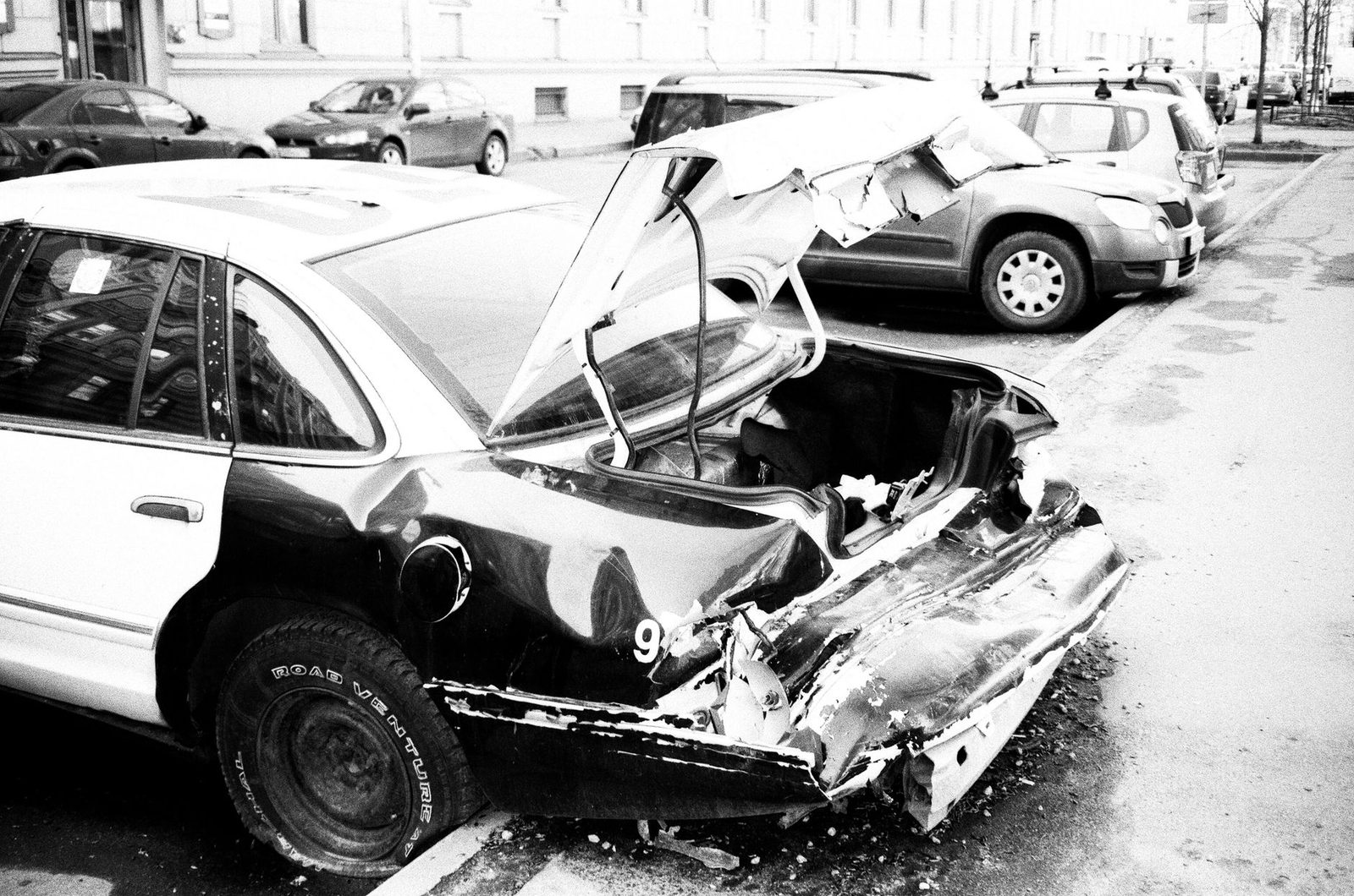 Black-and-white photo of a heavily damaged car on a city street.
