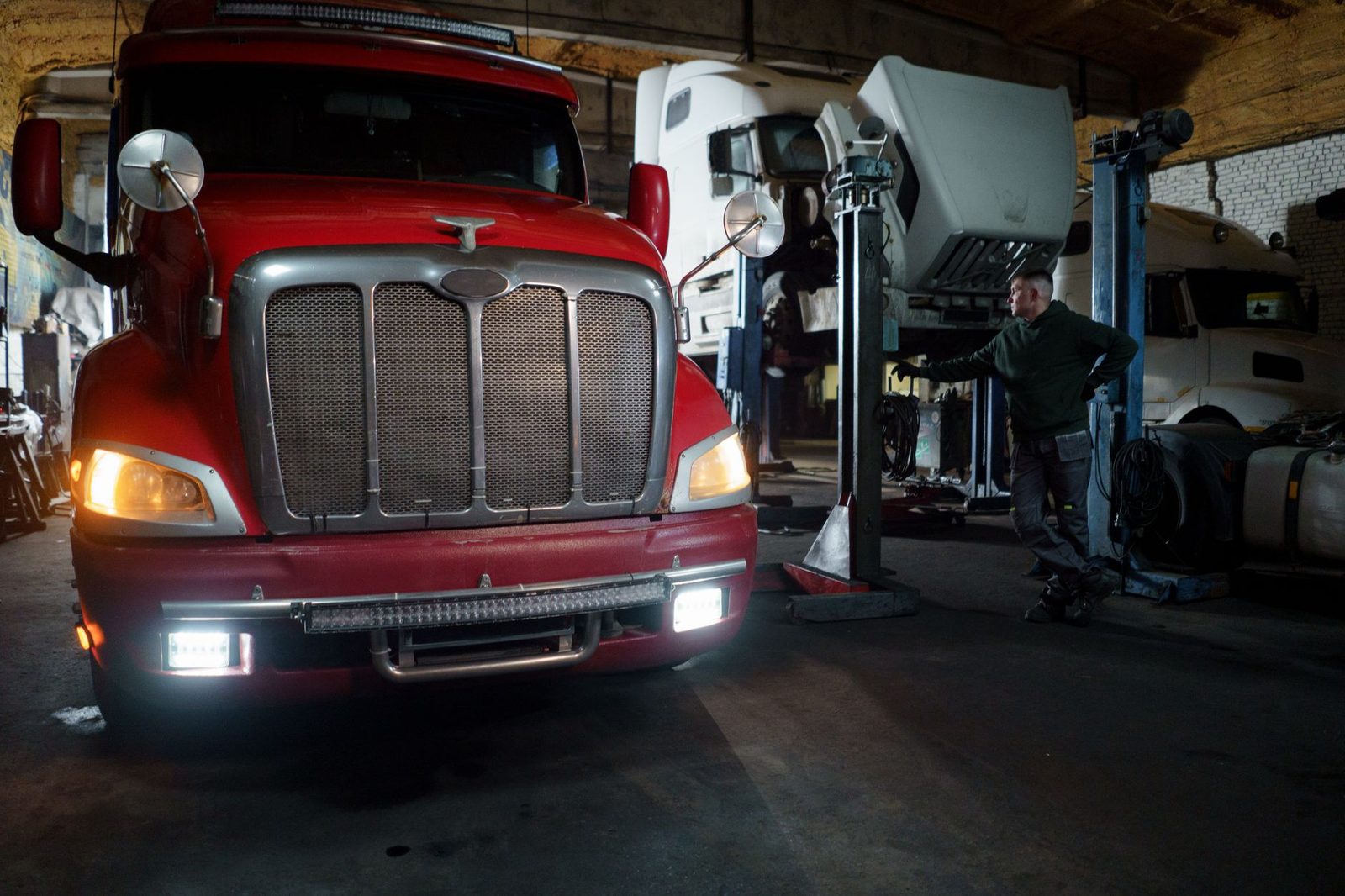 A mechanic works on a red truck in a garage, with other vehicles raised on lifts.