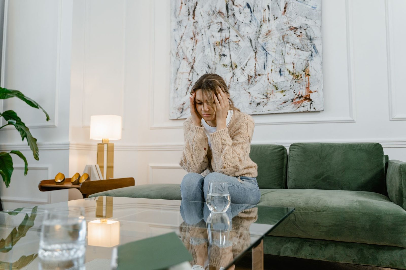 A woman sits on a sofa in a modern living room, looking distressed with her head in her hands.