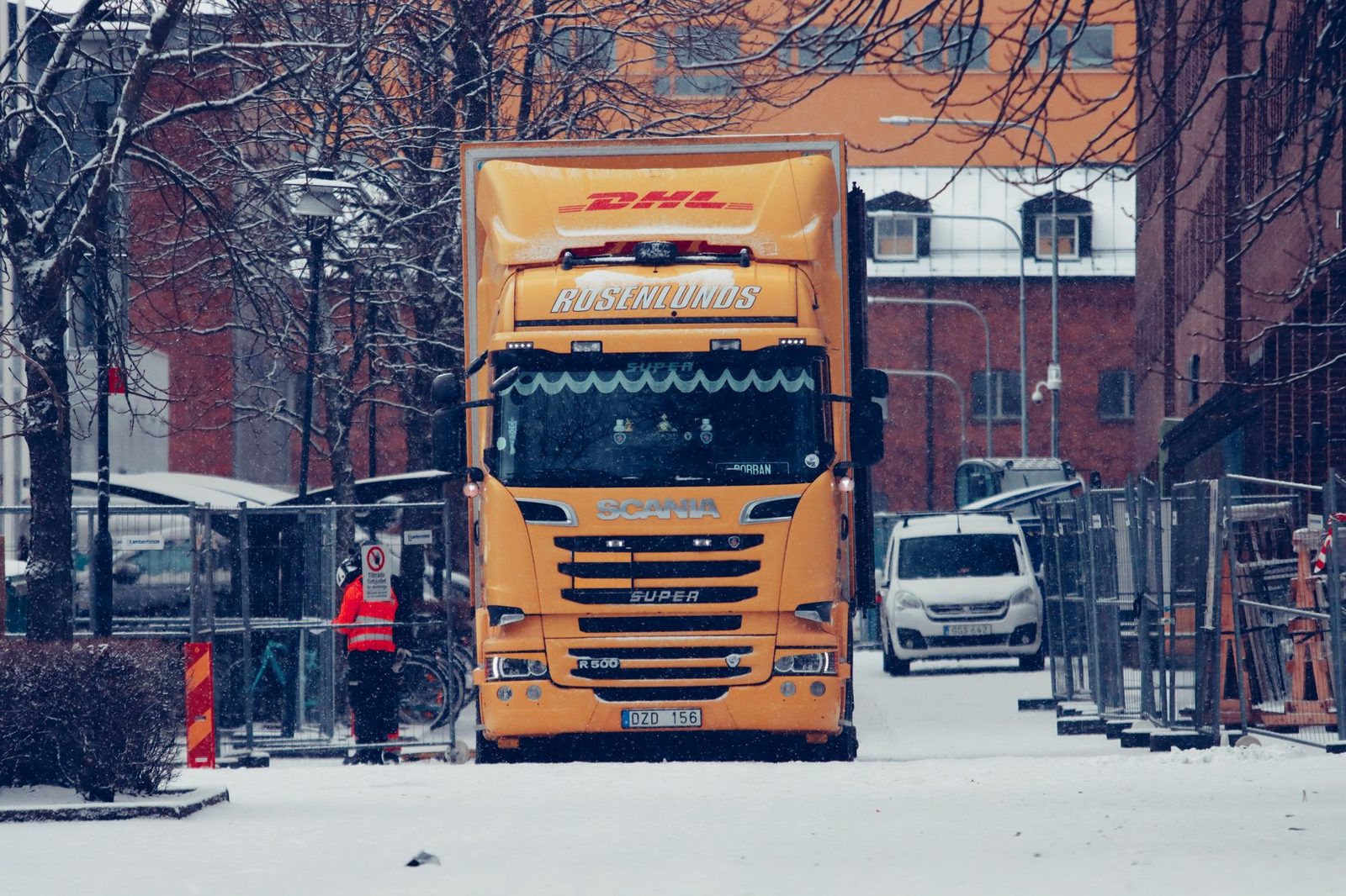 A yellow truck driving down a snow-covered city street lined with tall buildings.