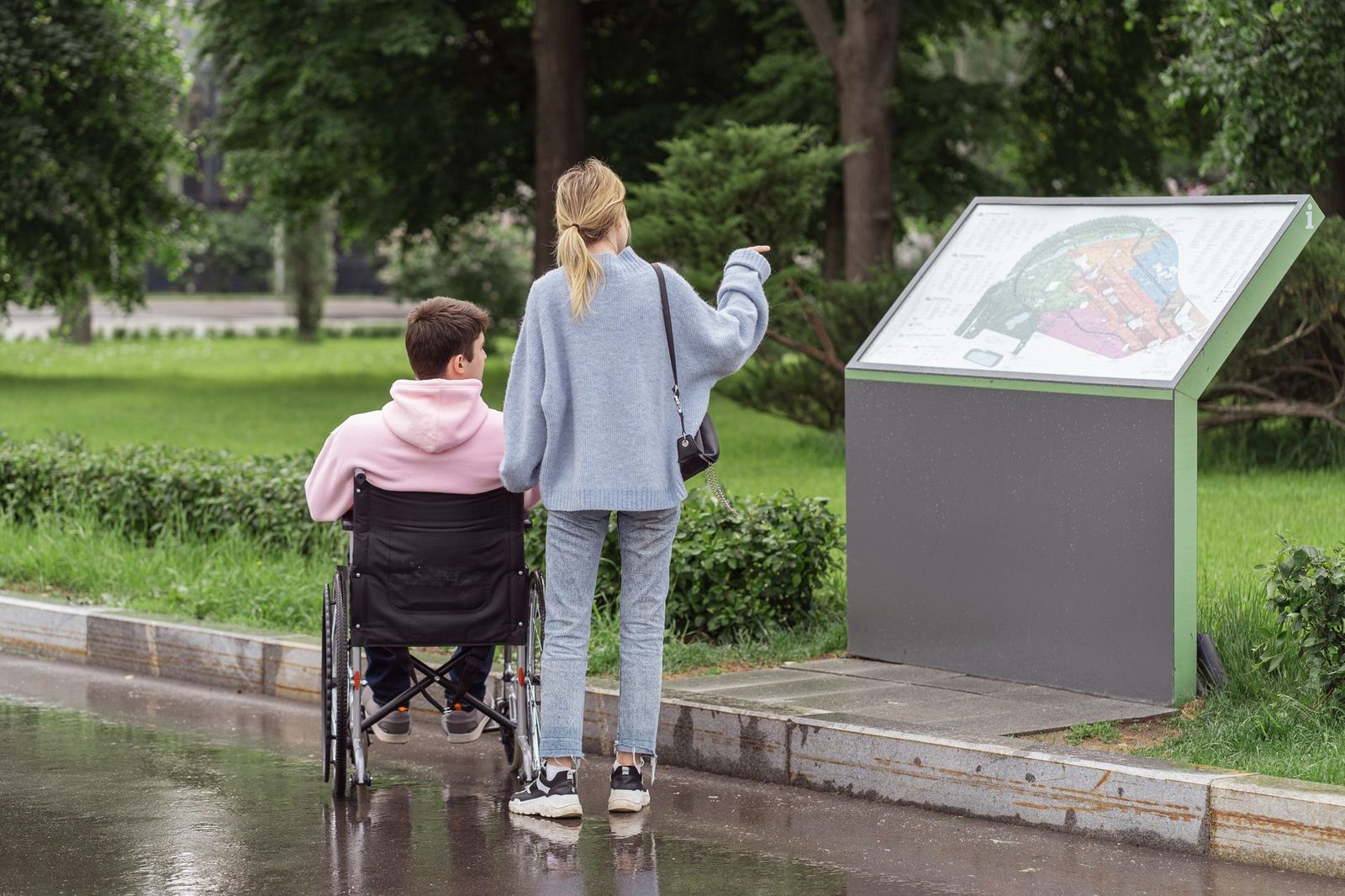 A young couple studies a park map on a rainy day, one partner seated in a wheelchair.