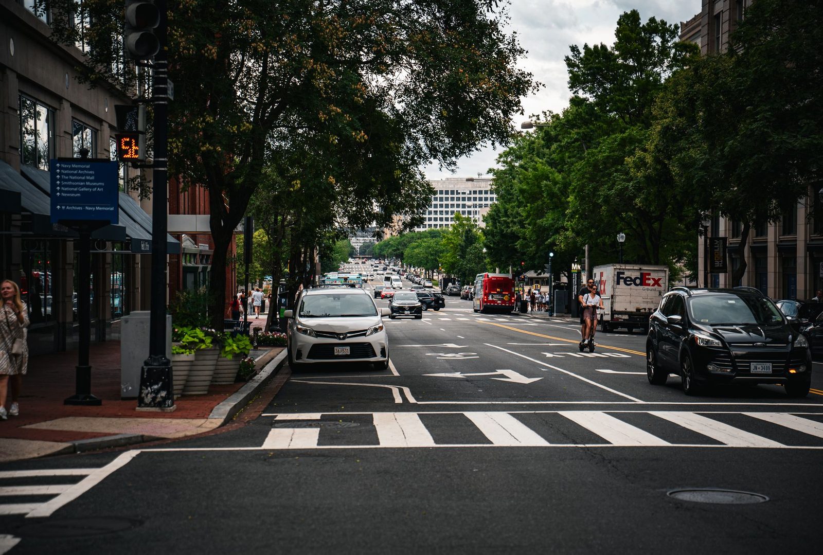 Busy city street lined with trees, with cars and pedestrians moving through an urban scene.