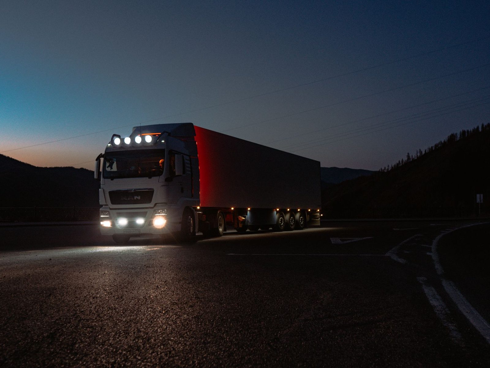 Heavy-duty truck driving on a mountain road at dusk with headlights on.