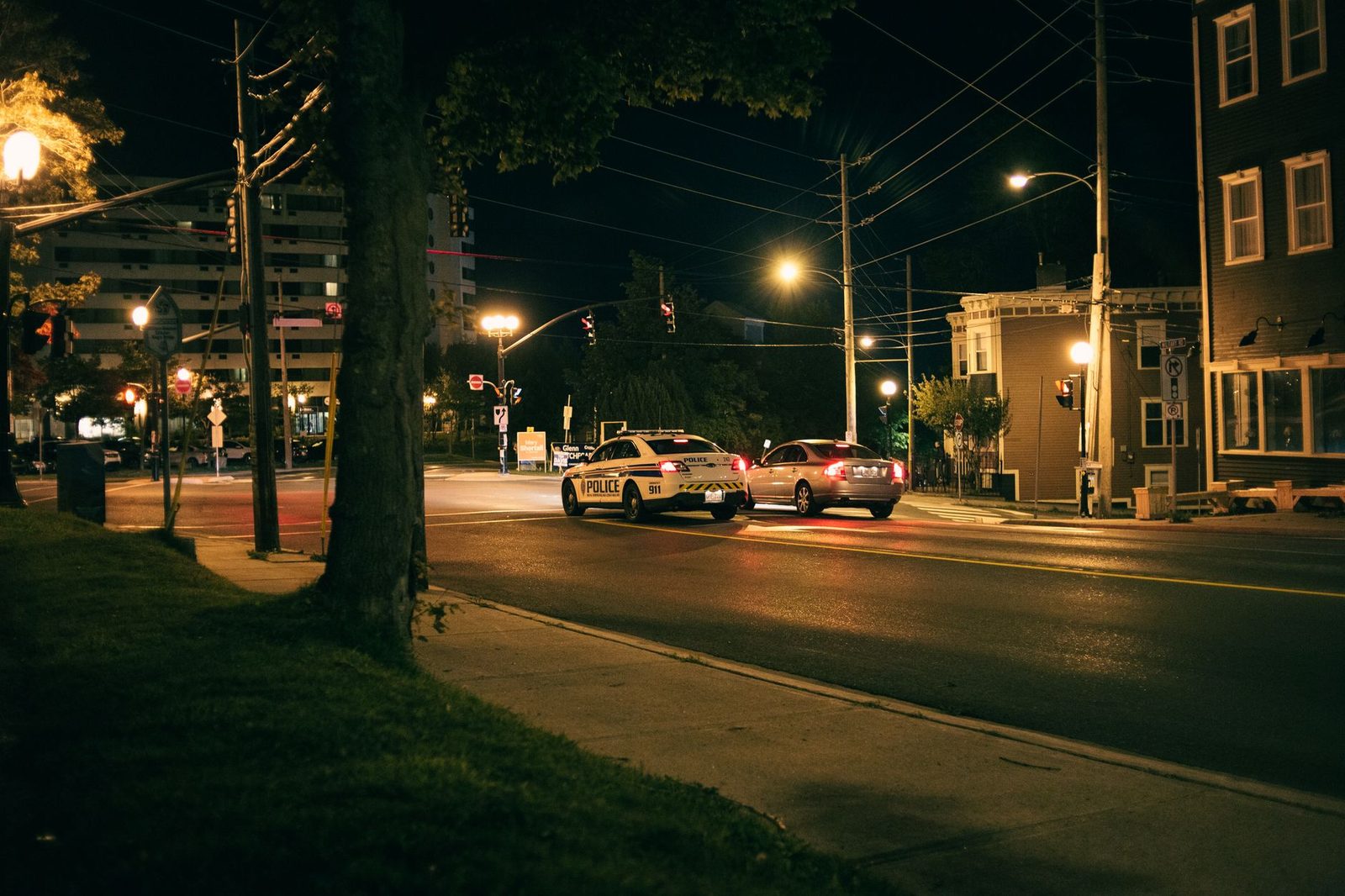 Nighttime city street scene with a police car and another vehicle lit by street lights.