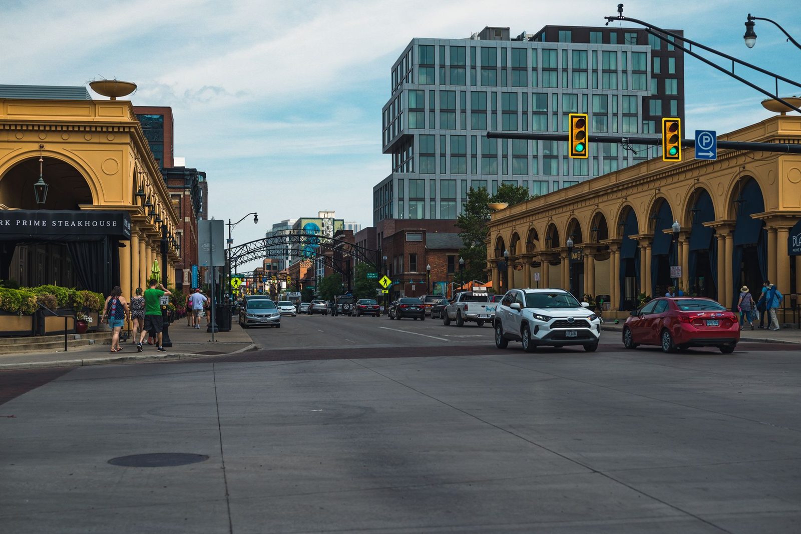 Pedestrians cross a busy city intersection on a clear day, with cars and tall buildings lining the street.