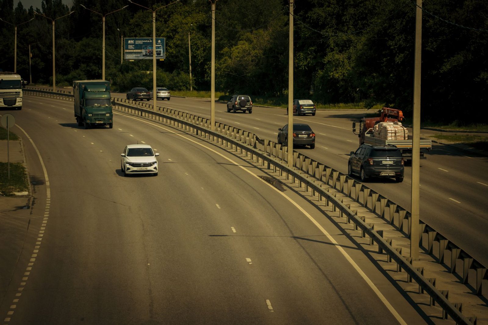 Several cars travel along a wide, sunny highway lined with trees.