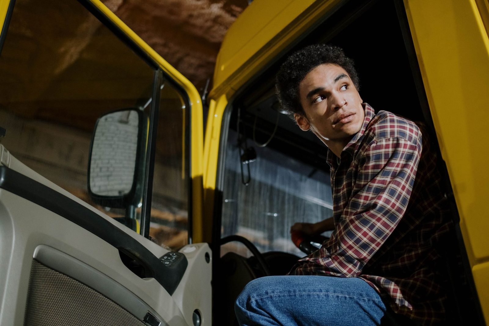 Young man in a plaid shirt sits inside a truck cab at night, looking out the window.
